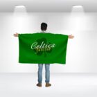 Person standing with their back to the camera, wearing a large green cloak with Celtics fanclub and a shamrock design printed on it. The room is well-lit with two overhead lights.