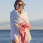A person wearing sunglasses is standing by the sea wrapped in a white and red fringed shawl. The sky is clear and the sunlight creates a soft, calm atmosphere.