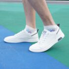 Close-up of a person wearing white sneakers with a unique design on a green and blue surface. The shoes have a pull tab at the heel and a logo on the side. The person is also wearing white socks with a colorful logo.