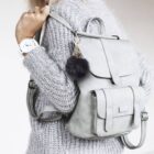 Person wearing a gray knit sweater, carrying a light gray backpack with a black fuzzy pom-pom keychain. The person has a white watch on their wrist and is facing away from the camera.