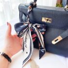 A person with red nail polish holds a black leather handbag with gold hardware. The bags handle is adorned with a tied, patterned navy scarf. The person is wearing a black wristwatch. The background features a white curtain and soft, natural light.