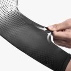 Close-up of a hand pulling the fabric of a black and white gradient sleeve, highlighting its elasticity. The fabric transitions from black to white with a dotted pattern. Two white arrows indicate stretching tension.