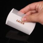 A hand holding a white comb with fine teeth against a black background. The word CAIXA is printed on the comb in brown letters.