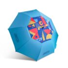 A blue umbrella with colorful geometric patterns, including circles, triangles, and rectangles, on the underside. The outer canopy is plain blue with Ogio logos on alternating panels.