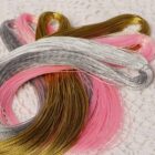 Colorful strands of metallic beads in silver, gold, pink, and copper are coiled neatly on a white lace surface. The beads shimmer in the light, creating a vibrant and rich display.
