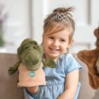 A young girl with a tiara smiles while holding a green dinosaur hand puppet, sitting on a couch. She is wearing a blue dress and there is another partially visible puppet in the foreground.