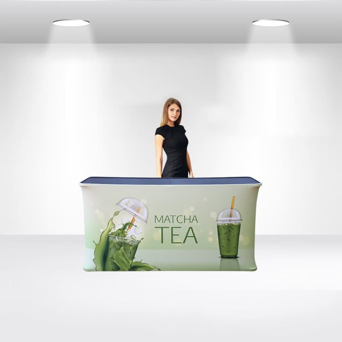 A person stands behind a promotional counter displaying a matcha tea advertisement. The counter features images of a matcha drink being poured and a cup with a straw. The background is a minimalist setting with white walls and ceiling lights.