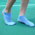 Person standing on a green mat wearing light blue socks with red non-slip dots on the soles. The socks have a logo with a blue circle and some text on the side. The persons legs are visible from the knees down.