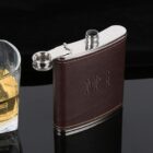 A dark leather hip flask with a metal cap is displayed next to a glass of whiskey on a glossy black surface. The flask has an engraved design and the cap is unscrewed, resting against it.