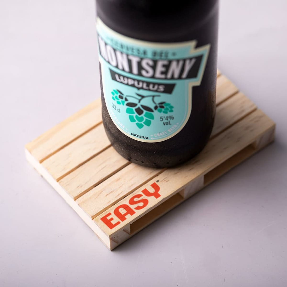 A bottle of beer labeled Montseny Lupulus rests on a wooden coaster with the word EASY printed in orange. The coaster features a slatted design, and the bottle is partially visible, displaying its green label with a hop graphic.
