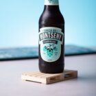 A dark glass bottle of Cerveza del Montseny Lupulus sits on a wooden coaster. The label is turquoise with an illustration of hop cones. The background is a gradient of light blue, and condensation is visible on the bottle.