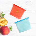 Two reusable silicone food bags, one red and one blue, are placed on a white wooden surface. A halved peach and a whole peach with leaves are next to the bags.