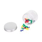 A clear plastic jar tipped over, spilling colorful push pins (red, green, blue, yellow, white) onto a white surface. The lid is placed beside the jar.