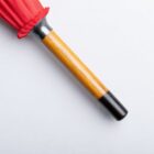 A wooden-handled, closed red umbrella is placed on a white surface. The handle is smooth with a black tip at the end. The folded fabric of the umbrella is tightly wrapped near the top of the handle.