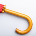 Close-up of a wooden umbrella handle with a curved end, attached to a folded red umbrella. The handle lies on a white surface.