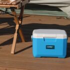 A blue and white cooler with a Cumbria logo is on a wooden deck next to a foldable wooden table. A large green fabric, possibly part of a tent or cover, is partially visible in the background.