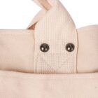 Close-up of a beige canvas bag with a textured fabric handle. Two metal rivets are visible, attaching the handle to the bag. The fabric has a natural, woven appearance.