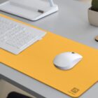 A white keyboard and mouse are placed on a vibrant yellow desk mat atop a gray desk. A white organizer and a potted plant are visible in the background. The desk mat has a logo in the bottom right corner.