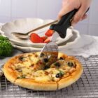 A hand uses a cutter to slice a freshly baked pizza topped with olives, mushrooms, and cheese. The pizza rests on a cooling rack. In the background, there are two plates with a spoon, a fork, and a few strawberries nearby.