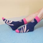 Two legs wearing blue socks with pink toe and heel accents, featuring abstract leaf patterns. The person is resting on a light blue floor against a cream-colored wall.