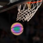 A basketball with a reflective, multicolored surface approaches the hoop, suspended in a mid-air moment just before passing through the white net, set against a dark background.
