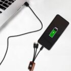 A smartphone connected to a laptop via a charging cable shows a green battery icon with a lightning bolt, indicating charging. The cable has multiple connectors, including a USB-C and micro USB, with a small orange adapter.