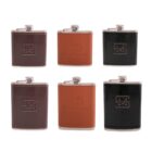 Six metal hip flasks with leather covers are displayed in two rows. The top row shows two black flasks with embossed logos on the leather and one brown flask. The bottom row shows one dark brown flask, one light brown with a logo, and one black flask.