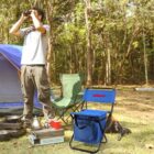 A person stands near a campsite, looking through binoculars. Theres a blue and black folding chair with a built-in cooler, a green chair, a tent, and camping gear, including a portable stove and cooking items, scattered on the grassy ground.