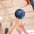 A basketball player reaches up to a colorful, iridescent basketball as it approaches the hoop. Multiple hands are visible, emphasizing the action and intensity in an indoor court setting.