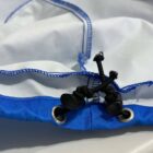 Close-up of a blue and white fabric, possibly a drawstring bag, with two black elastic cords threaded through metal eyelets. The cords are secured with plastic toggles and knotted. The fabric features visible blue stitching along the edges.