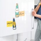 A person holding the handle of a white fridge door. The fridge has two bottle-shaped magnets and sticky notes, one reading URGENT and the other text message here. The person is wearing a blue sleeve and black pants.