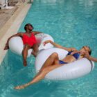 Two people relax on inflatable rings in a swimming pool. One person in a red swimsuit enjoys the sun, while the other in a blue patterned swimsuit leans back with arms extended, both floating peacefully in the calm water.