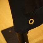 Close-up of a black fabric banner featuring a brass grommet. The banner is attached to a vertical black metal pole, set against a light brown wooden surface.