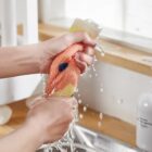 Two hands are squeezing a novelty sponge shaped like a muscle, with water pouring over it. The sponge has a red, fleshy appearance with a black spot. The scene takes place over a kitchen sink.