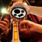 A hand holding a circular black bottle opener above a brown bottle, ready to open it. In the background, several out-of-focus hands are holding up bottles in a celebratory gesture. The setting suggests a festive atmosphere.