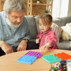 A bearded man and a young girl sitting at a table with colorful pop-it toys. The girl is focused on playing, while the man watches. The setting is a cozy room with shelves and a couch in the background.