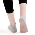 A person wearing light gray grip socks with orange traction pads on the soles. They are standing on a light surface, and only their lower legs, covered with black leggings, are visible from the back.