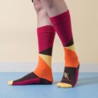 Person wearing colorful socks with geometric patterns in orange, yellow, pink, and brown tones standing on a blue floor with a beige background.