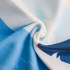 Close-up of a soft, mixed-textured fabric with a swirl pattern. Predominantly light blue and white, featuring darker blue sections. The fabric appears silky with subtle design elements, creating a gentle, flowing appearance.