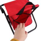 A hand wearing a yellow sleeve is seen opening a red foldable stool with a zippered compartment on a black metal frame. The stool doubles as a storage bag.