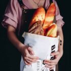 Person holding a cloth bag containing several loaves of bread, including baguettes and a rustic loaf. They are wearing a pink shirt and an apron, set against a dark background.