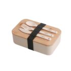A rectangular lunch box with a bamboo lid secured by a black strap. Three utensils—a fork, spoon, and knife—are on top, held in place by the strap. The container is cream-colored and minimalist in design.