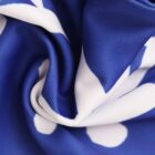 Close-up of a blue fabric with white polka dots, artistically twisted and folded. The fabric appears smooth and silky, creating loops and curves with contrast between the rich blue and bright white patterns.