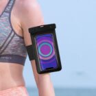 A person wearing a colorful sports bra uses a black armband to hold a smartphone. The phone displays a bright circular pattern in blue, purple, and green. The background is a clear blue sky.