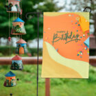 A garden flag with Happy Birthday written in green on an orange and yellow background, adorned with colorful confetti, is displayed outdoors. A row of decorative, mini house-shaped wind chimes hangs nearby, with greenery in the background.