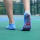 A person walking on a green surface wearing light blue socks with red grip patterns on the sole. The background shows an outdoor area with a blue and red floor.