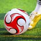 A close-up of a soccer ball featuring a red and black design being kicked by a person wearing golden shoes. The ball is on a vibrant green grass field.