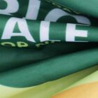 Close-up of a green and yellow fabric with partially visible white text. The material is slightly folded, creating shadows and highlights across the surface. The text includes the letters ALE.