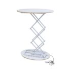 A round, adjustable white table with a scissor lift mechanism. The tabletop is supported by a collapsible metal frame on an oval base. An electrical cord extends from the base.