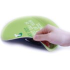 A hand lifting the edge of a flexible, green rubberized mat featuring white text. The mat appears to have a glossy finish, and part of the text on the visible top reads FIRST DAY OF YOUR and NESS.